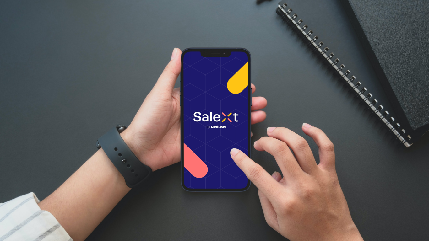 A person's hands hold and touch a smartphone displaying the Salext by Mediaset app logo screen, resting on a dark desk near a spiral notebook
