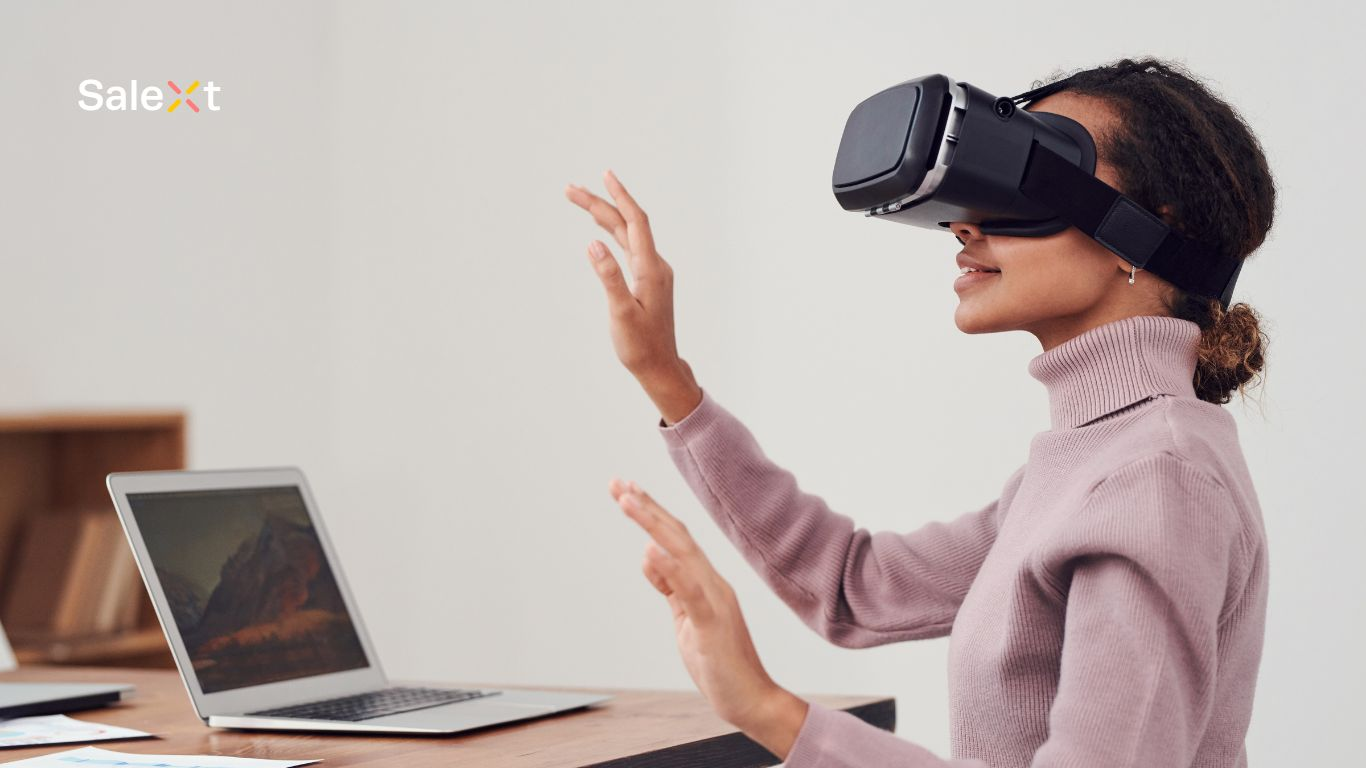 A woman wearing a VR headset gestures with both hands raised near a desk with an open laptop